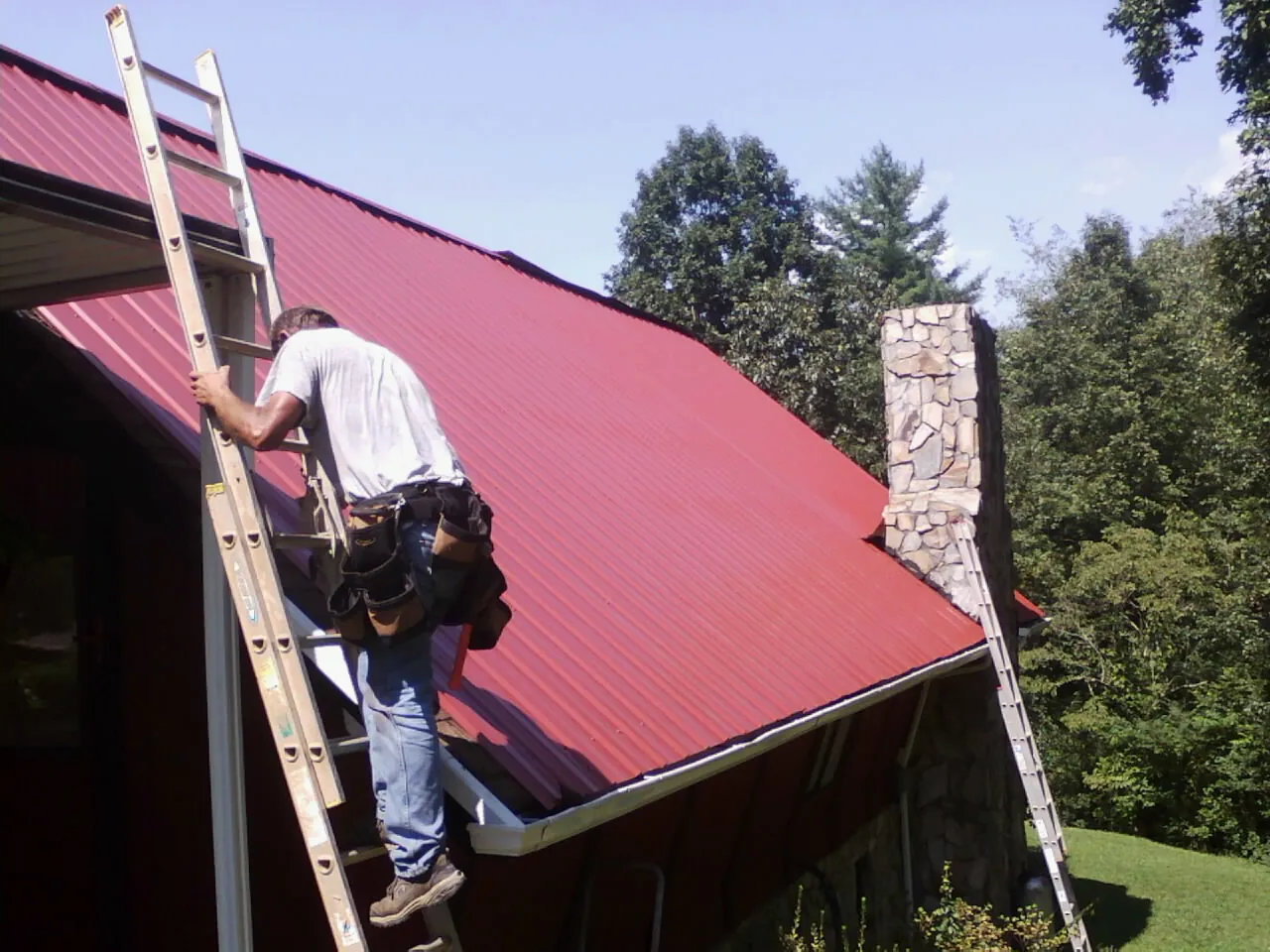 Roofer on a ladder beside a red metal roof and stone chimney — Soffit & Fascia Repair by Roofing Brothers in Kirksville